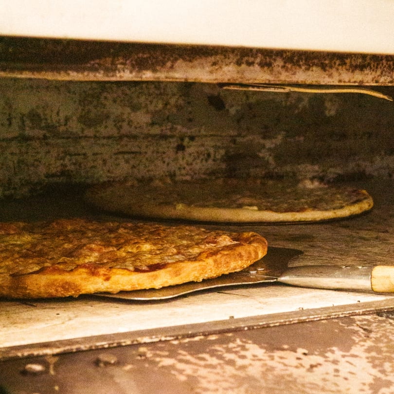 Two pizzas baking inside a rustic, well-used pizza oven, one on a metal peel.