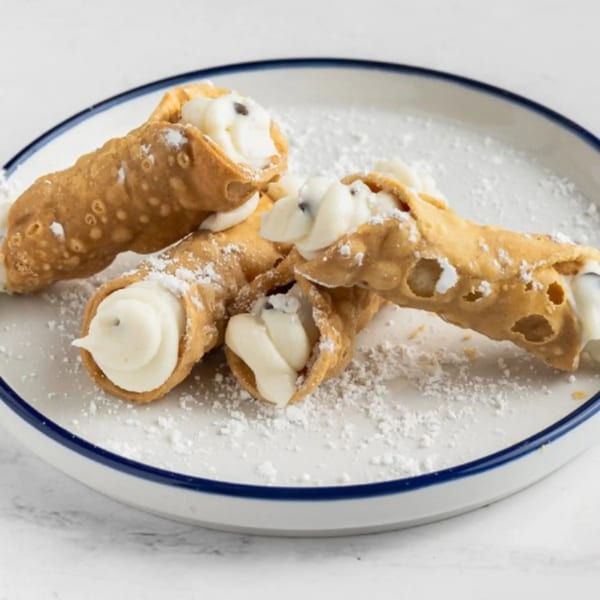 Four golden-brown cannoli filled with white cream and sprinkled with powdered sugar on a white plate.