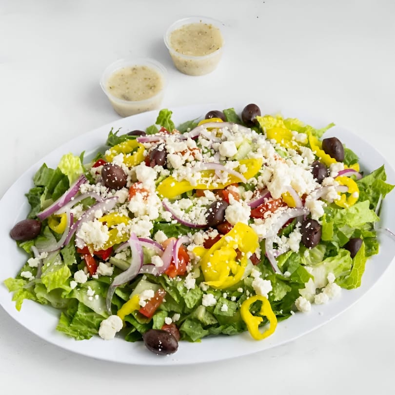 A large Big City Slices Greek salad with feta, olives, peppers, and dressing cups on a white plate.