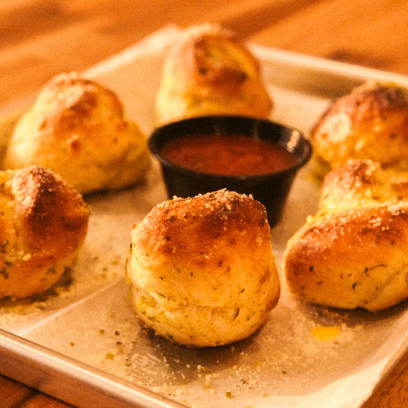 Golden-brown garlic knots with parmesan cheese and herbs, served with a side of marinara sauce.