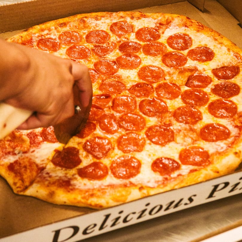 A hand uses a pizza cutter to slice a large pepperoni pizza in a Big City Slices box.