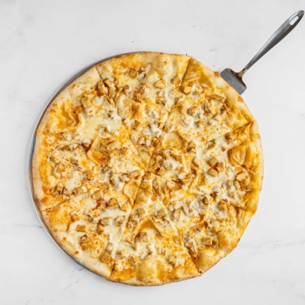 A whole Big City Slices chicken pizza, cut into slices, with a spatula on a white surface.