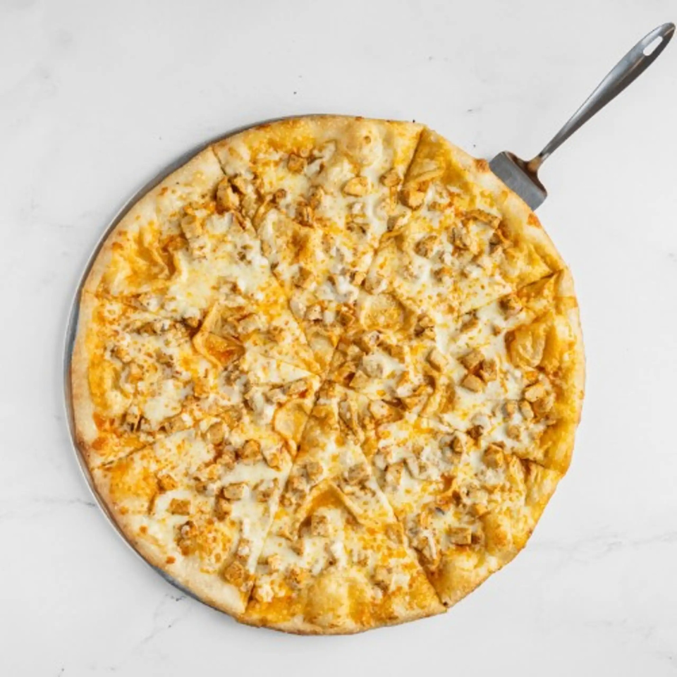 A whole Big City Slices chicken pizza, cut into slices, with a spatula on a white surface.